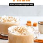 Pinterest image of two cups of Caramel Milk on a coaster surrounded by caramel candies, caramel syrup, and leaves.