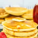 Pinterest image of a large stack of Cracker Barrel pancakes on a plate with a slice of butter and syrup on top and berries on the side, with more pancakes in the background.