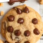 Pinterest image of an overhead shot of a chocolate chip cookie on a cooling rack with baking paper, cookie crumbs, chocolate chips, and two more cookies on the side.