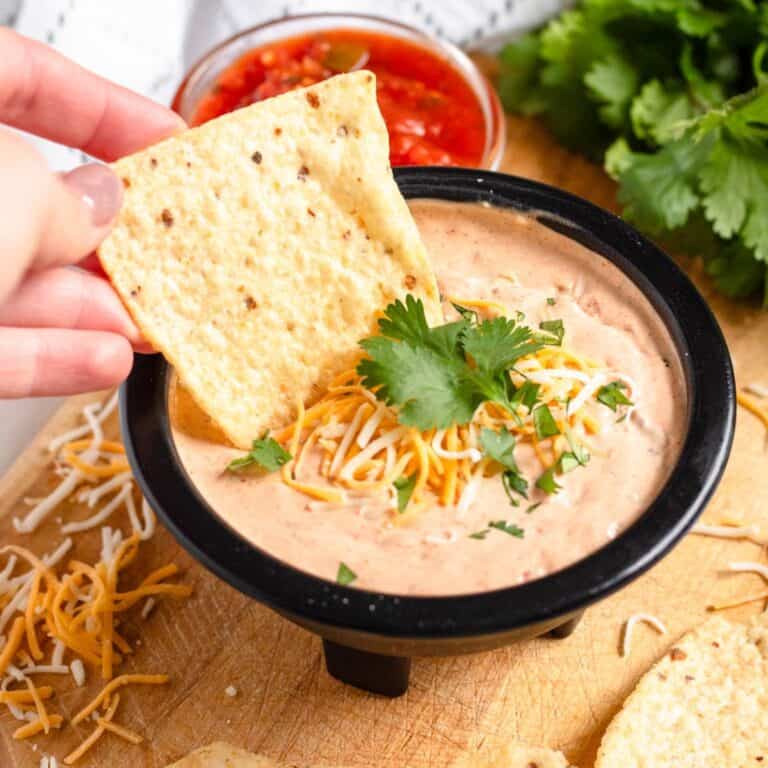Close up of a tortilla chip dunking into a small bowl of Mexicali dip with cilantro and shredded cheese garnished on top.
