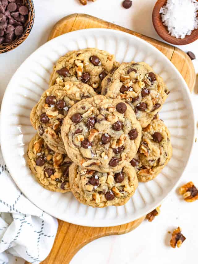 Top view of a stack of freshly baked chocolate chip and walnut cookies on a plate with walnuts and a small bowl of sea salt laying around the plate.