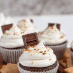 Pinterest image of s'mores cupcakes with marshmallow frosting on a serving board with chunks of Hershey chocolate bars and graham cracker pieces on the side.