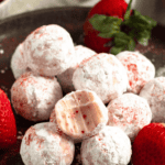 Pinterest image of a plate of strawberry truffles coated in powdered sugar and dusted with freeze-dried strawberry powder, with a bite taken out of one of the truffles showing the creamy strawberry white chocolate filling, surrounded by fresh strawberries.