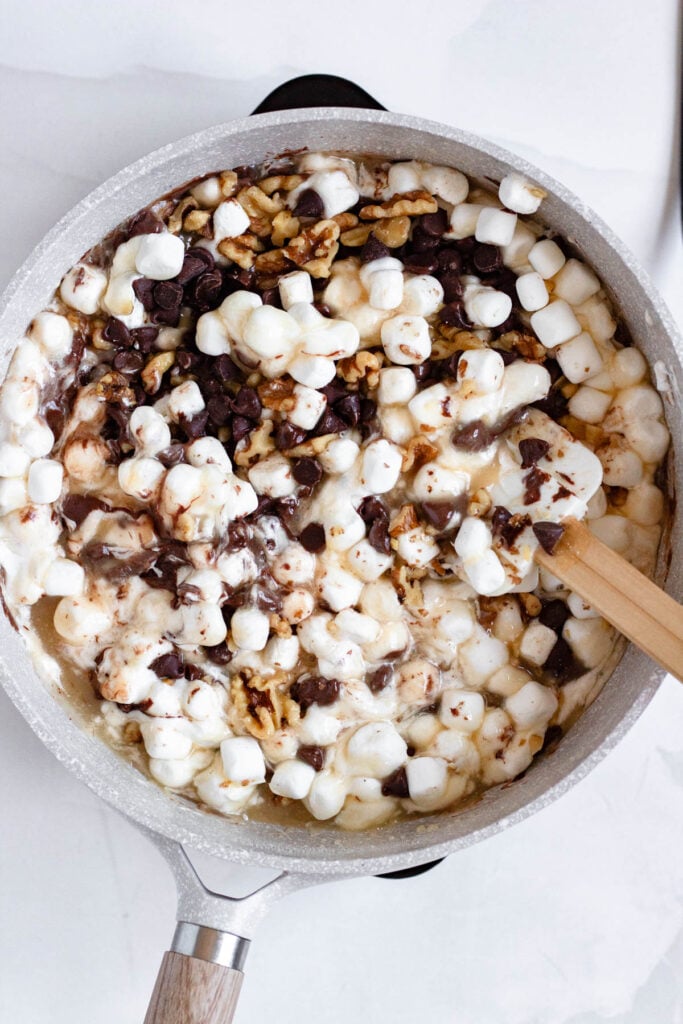A pot of melting marshmallows, chocolate chips and walnuts being stirred together with a wooden spatula.