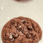 Pinterest image of a freshly baked double-chocolate chip cookie topped with sea salt sits on a metal baking sheet.