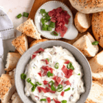 Pinterest image of a top view of creamy bagel dip topped with chopped green onions and deli meat, surrounded by bagel pieces.