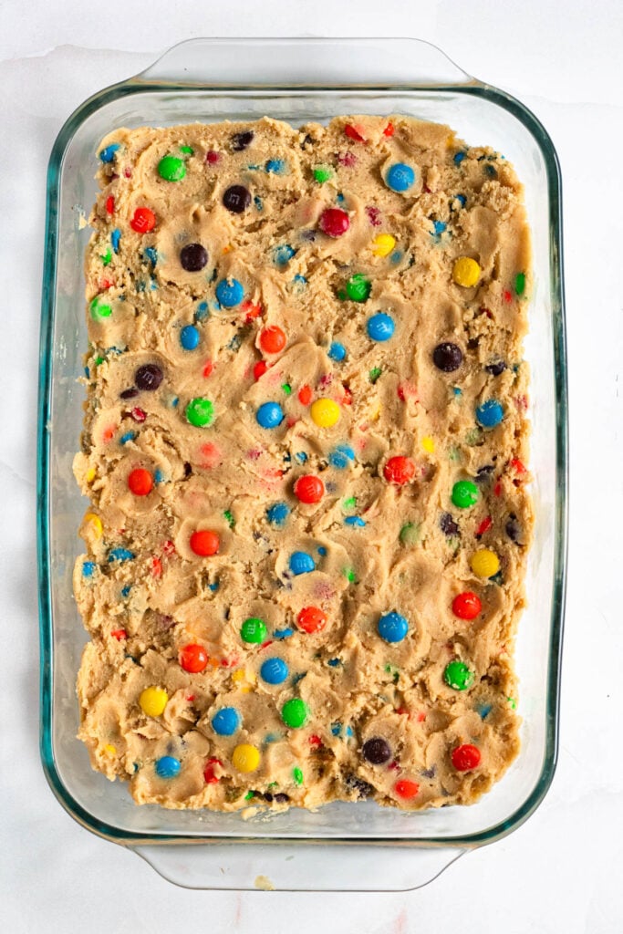 Unbaked M&M cookie dough spread in a glass baking dish.