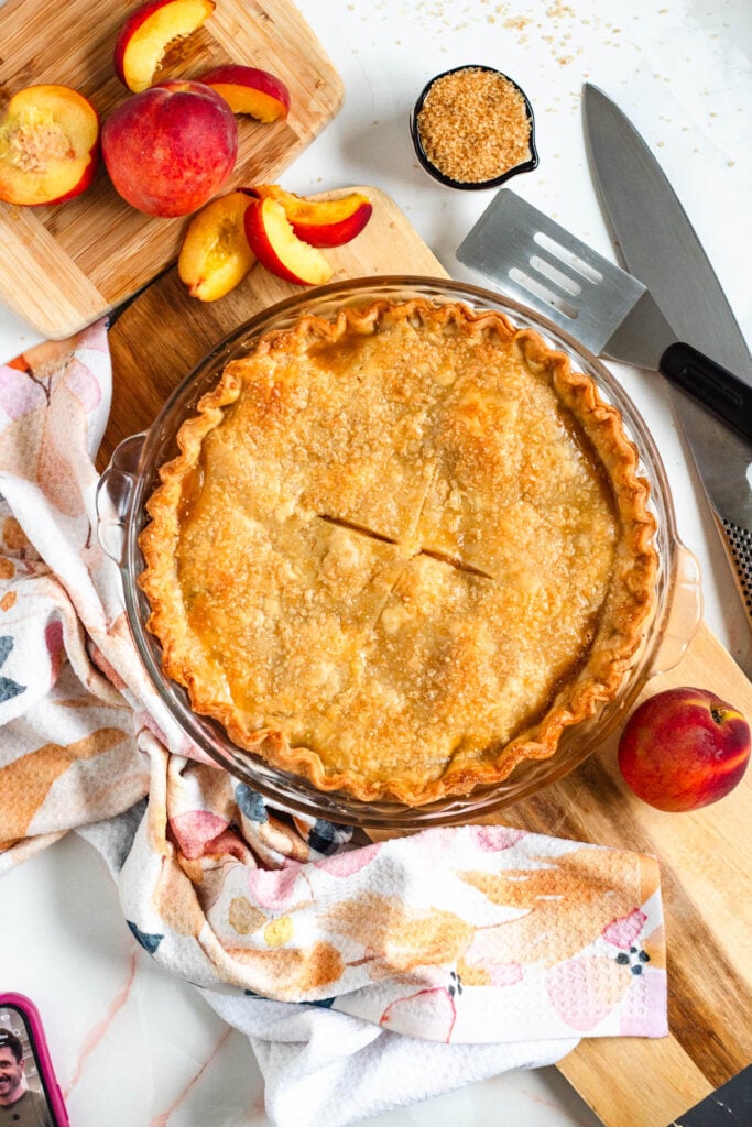 A golden peach pie rests on a wooden board, surrounded by peach slices.