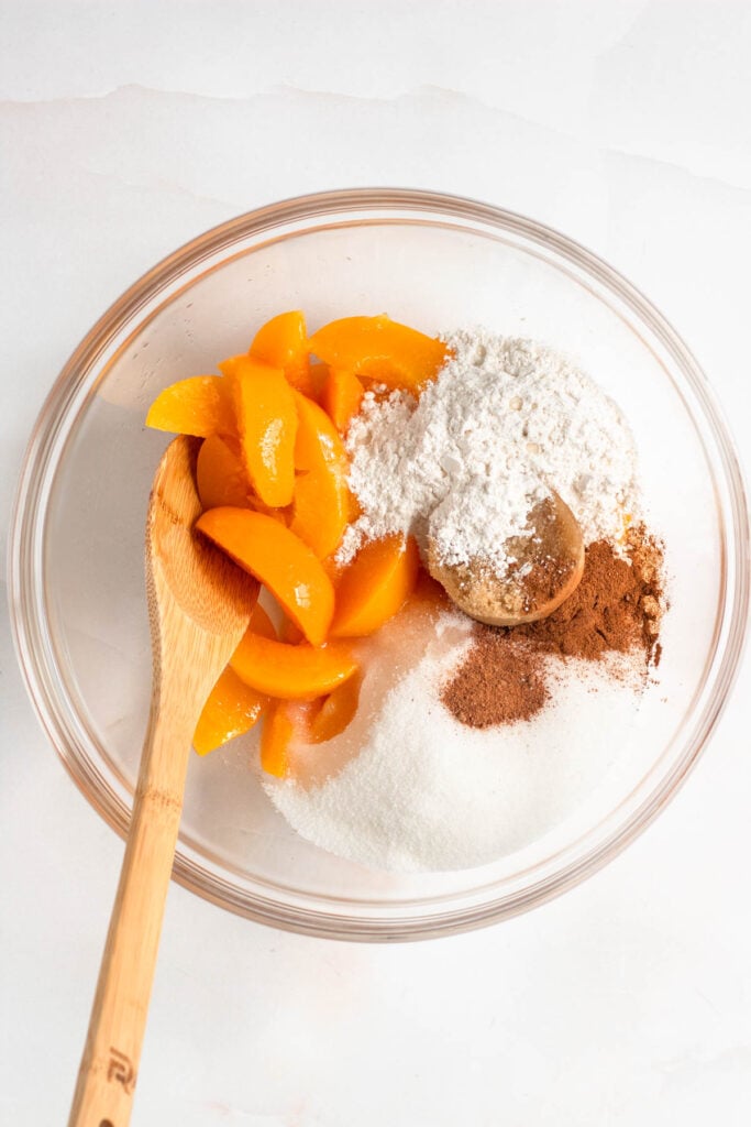 Drained peaches, granulated sugar, brown sugar, cornstarch, cinnamon, nutmeg, and lemon juice were added to a bowl, ready to be mixed.