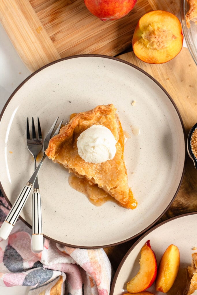 A slice of peach pie topped with creamy vanilla ice cream sits on a plate, with two forks resting beside it.