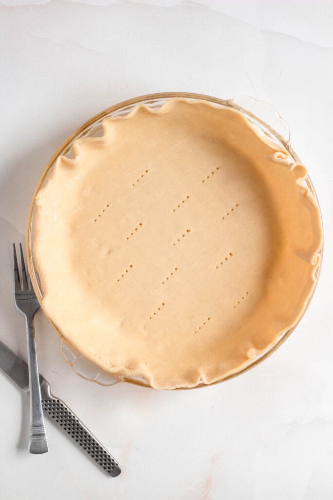 A sheet of pie dough is neatly rolled out into a pie dish, with fork marks pricked across the bottom.