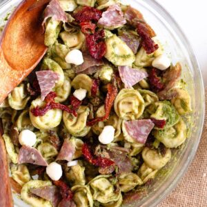 A vibrant pesto tortellini pasta salad loaded with sun-dried tomatoes, mozzarella pearls, and salami.