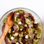 Pinterest image of an overhead shot of a bowl of pesto tortellini pasta salad that is well-coated in green pesto and mixed with sun-dried tomatoes, mozzarella, and salami, with two wooden serving spoons.