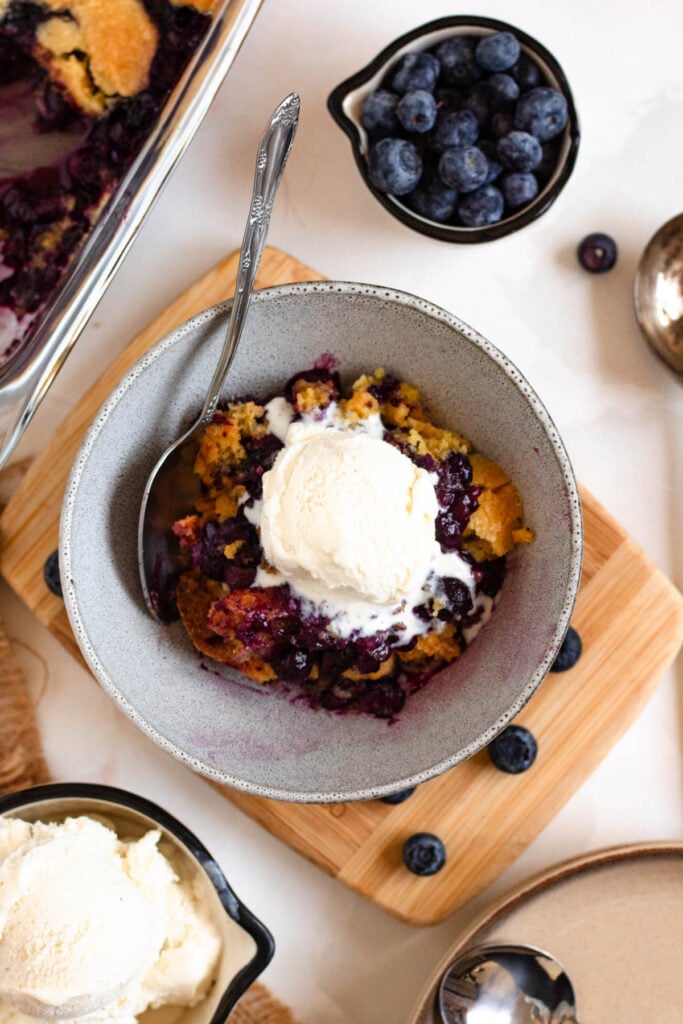 A bowl of warm, gooey 3-ingredient blueberry cobbler topped with melting ice cream, surrounded by extra bowls of ice cream and fresh blueberries on the side.