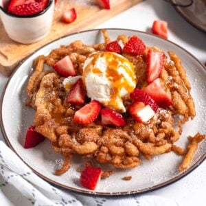 A golden, crispy funnel cake topped with fresh strawberries, a scoop of vanilla ice cream, and a drizzle of caramel sauce.