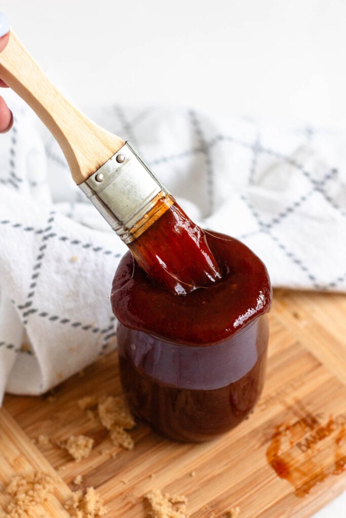 Thick barbecue sauce coats a basting brush over a jar, with brown sugar scattered on a wooden board.