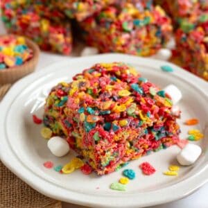 A piece of Fruity Pebbles Rice Krispie Treat sits on a plate next to mini marshmallows, with the rest of the colorful batch in the background.