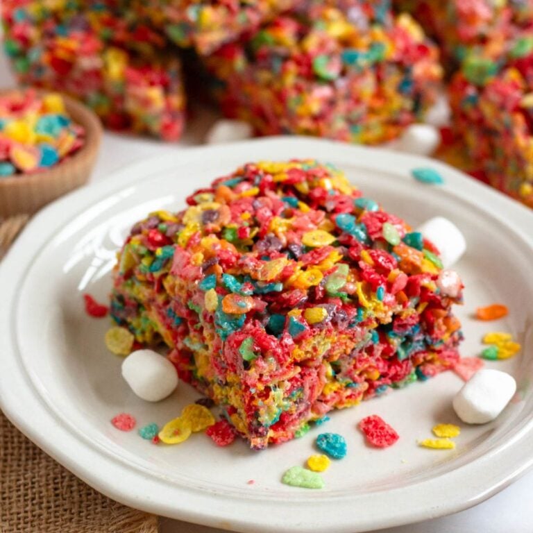 A piece of Fruity Pebbles Rice Krispie Treat sits on a plate next to mini marshmallows, with the rest of the colorful batch in the background.