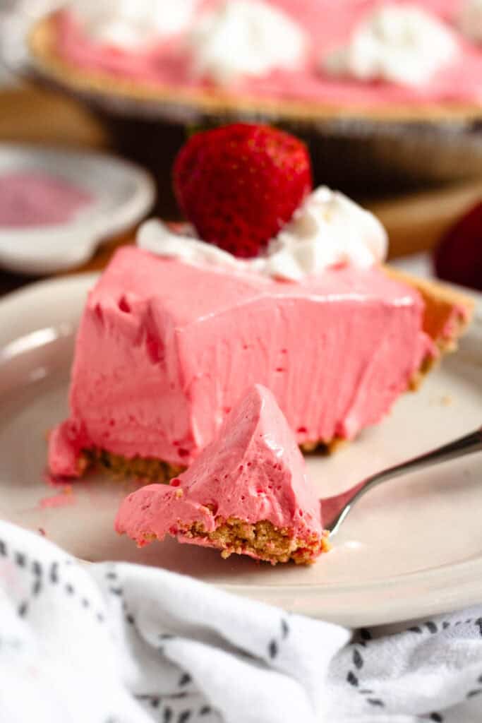 A slice of pink Kool-Aid pie, topped with whipped cream and a strawberry, with a fork lifting a bite-sized piece.