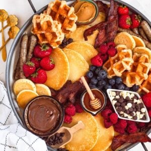 A breakfast charcuterie board filled with pancakes, waffles, bacon, sausages, fresh berries, and sweet toppings like Nutella, honey, and chocolate chips