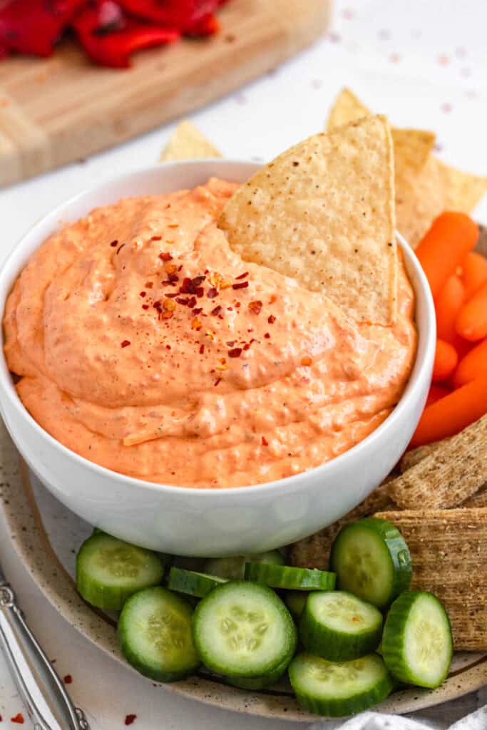 Creamy roasted red pepper dip served with tortilla chips, cucumbers, and veggies on a platter.