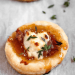 Pinterest image of a close-up shot of goat cheese puff pastry bites on parchment paper, with herbs on the side.