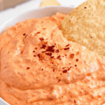 Pinterest image of a close-up shot of a tortilla chip dipped in the creamy roasted red pepper dip served with tortilla chips, cucumbers, and veggies on a platter.