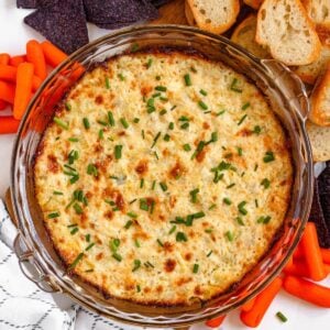A golden, bubbly hot artichoke dip topped with fresh chives is served with chips, crostini, and carrots.