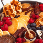 A Breakfast Charcuterie Board filled with pancakes, waffles, bacon, sausages, fresh berries, and sweet toppings like Nutella, honey, and chocolate chips