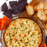 Pinterest image of an overhead shot of the golden, bubbly hot artichoke dip topped with fresh chives, served with chips, crostini, and carrots.