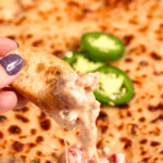 Pinterest image of a close-up shot of hand holding a toasted bread slice dipped into Sausage Cheese Dip with a melty, stretchy texture and sliced jalapeños in the background.