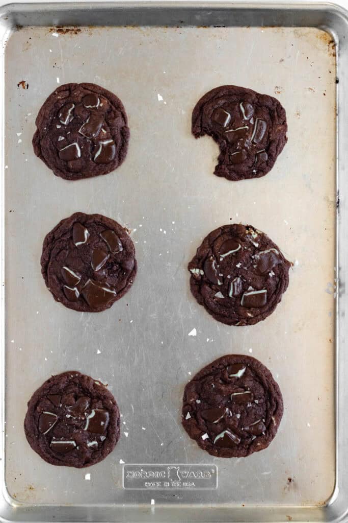 Six Andes mint chocolate cookies with melted chocolate chunks on a baking sheet, one partially eaten, topped with flaky sea salt.