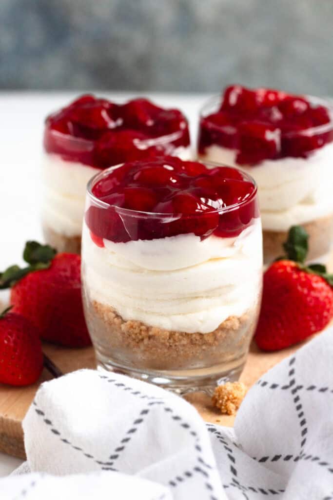 Three-layered no-bake cheesecake cups in clear glasses topped with strawberry pie filling.