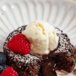 Pinterest image of the Molten Chocolate Lava Cake on a plate with a bite taken out, topped with vanilla ice cream and raspberry, with fresh berries on the side and melted chocolate oozing out.