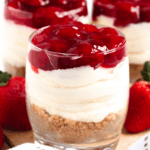 Pinterest image of three-layered, no-bake cheesecake cups in clear glasses topped with strawberry pie filling, with fresh strawberries beside them.