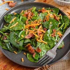 Spinach salad on a plate with cheddar cheese and bacon, and a fork resting on the plate.