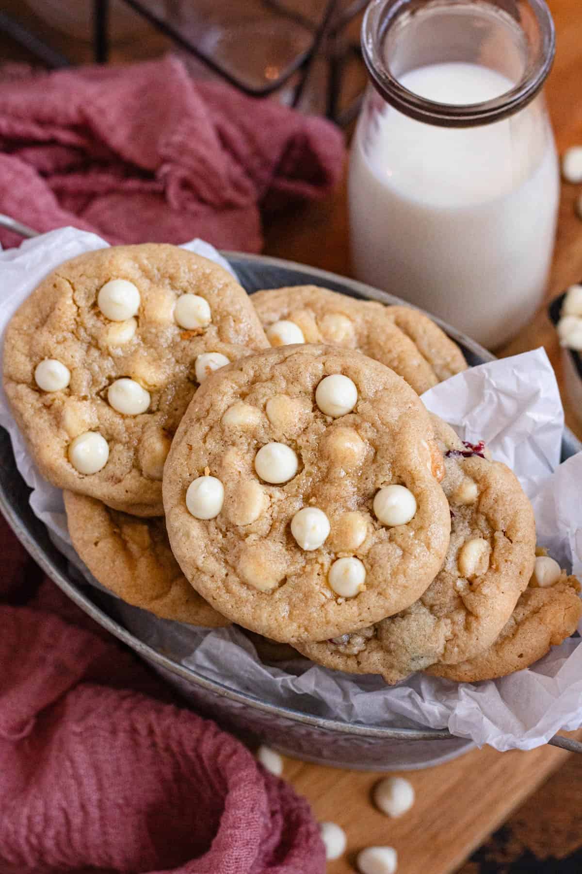 Freshly baked white chocolate chip cookies in a silver tin next to a glass of milk