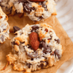 Pinterest image of a close-up shot of baked Almond Joy Cookies with coconut, chocolate chips, and whole almonds on a parchment-lined plate.