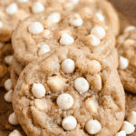 Pinterest image of a close-up shot of a stack of white chocolate chip cookies resting on brown parchment paper with white chocolate chips surrounding it.